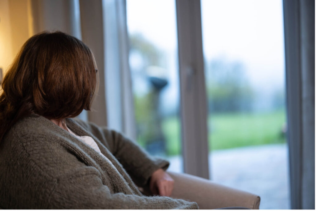 Frau mit braunen Haaren und grauem Pullover sitzt auf einem Stuhl und blickt aus einem Fenster auf eine grüne Landschaft