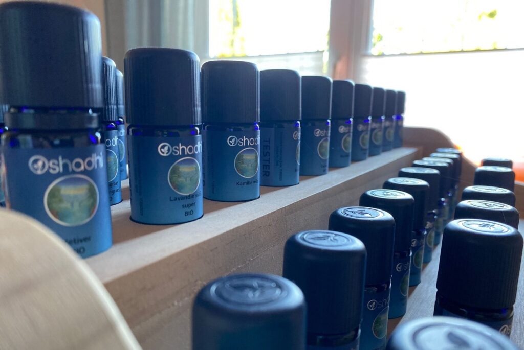 Reihe von blauen ätherischen Ölfläschchen der Marke shadhi auf einem Holzregal vor einem Fenster