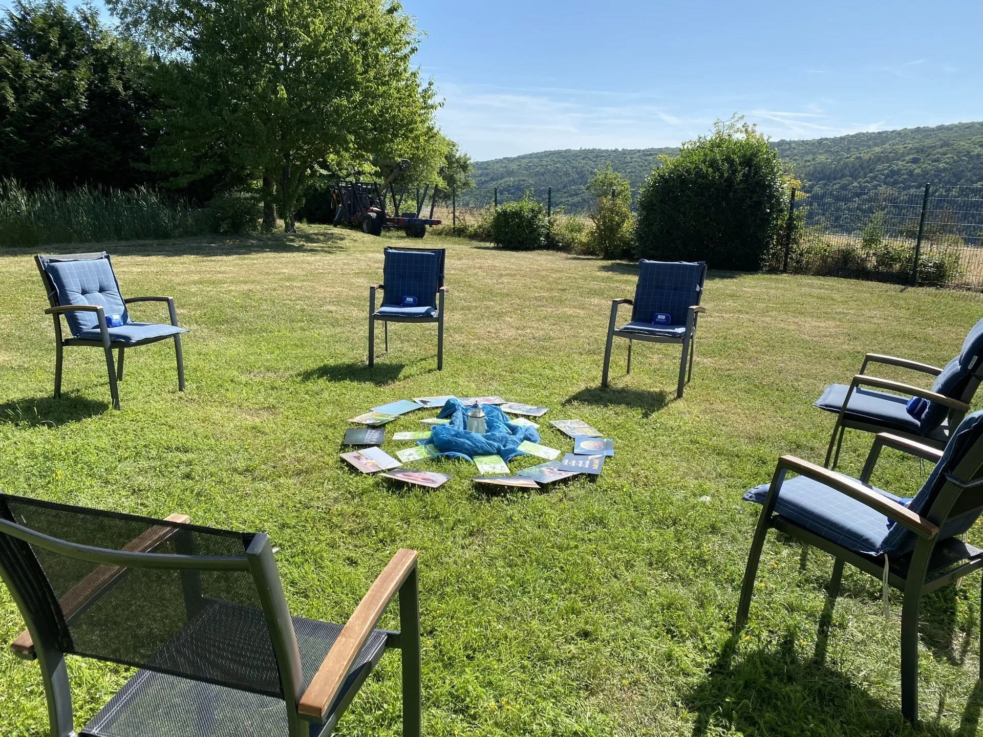 Stuhlkreis mit blauen Sitzkissen auf einer Wiese, in der Mitte Bücher und ein Teelicht auf blauem Tuch, umgeben von Bäumen und Hügeln im Hintergrund