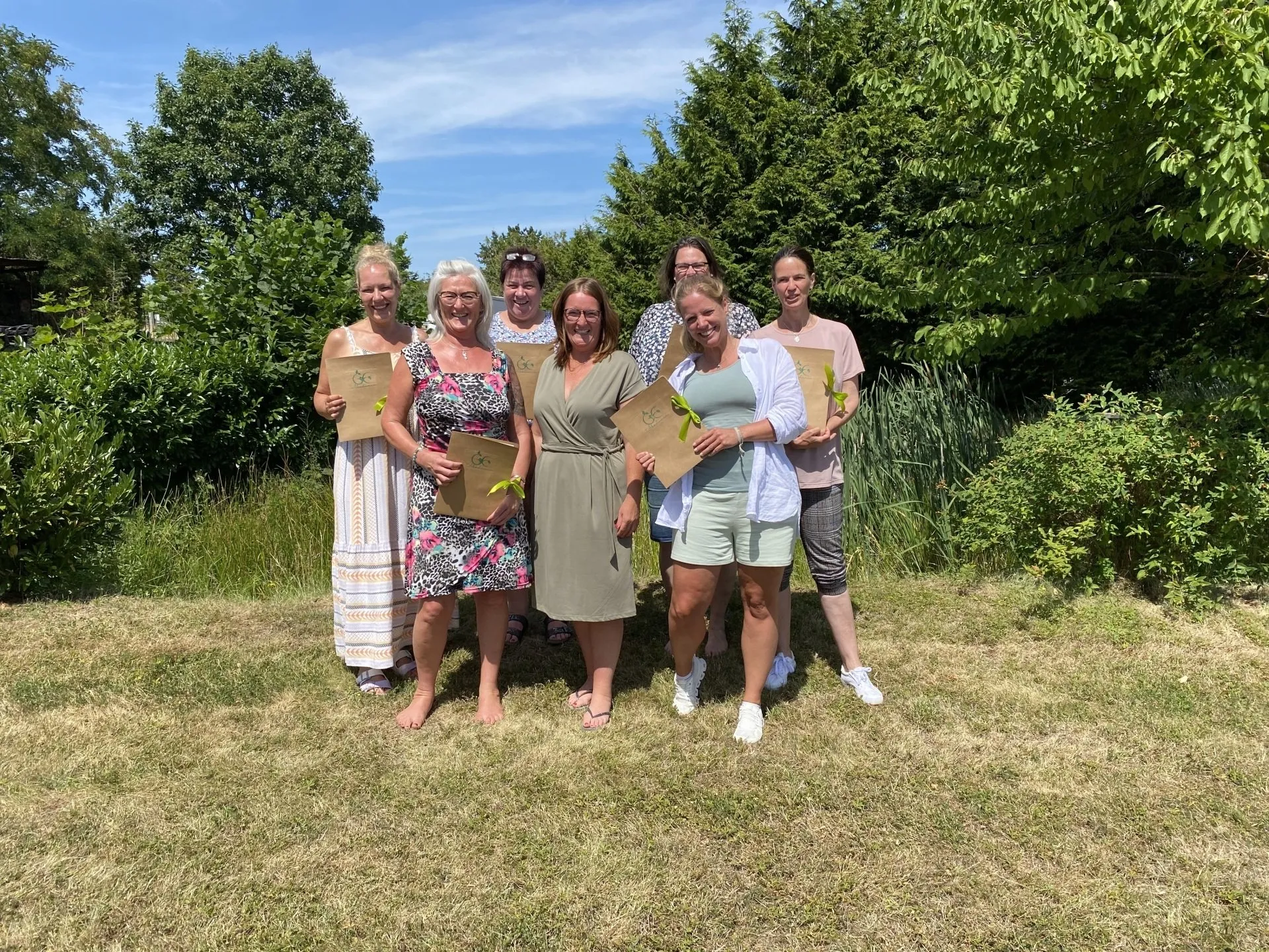 Sieben Frauen stehen auf einer Wiese vor Büschen, halten Zertifikate mit grüner Schleife und lächeln in die Kamera