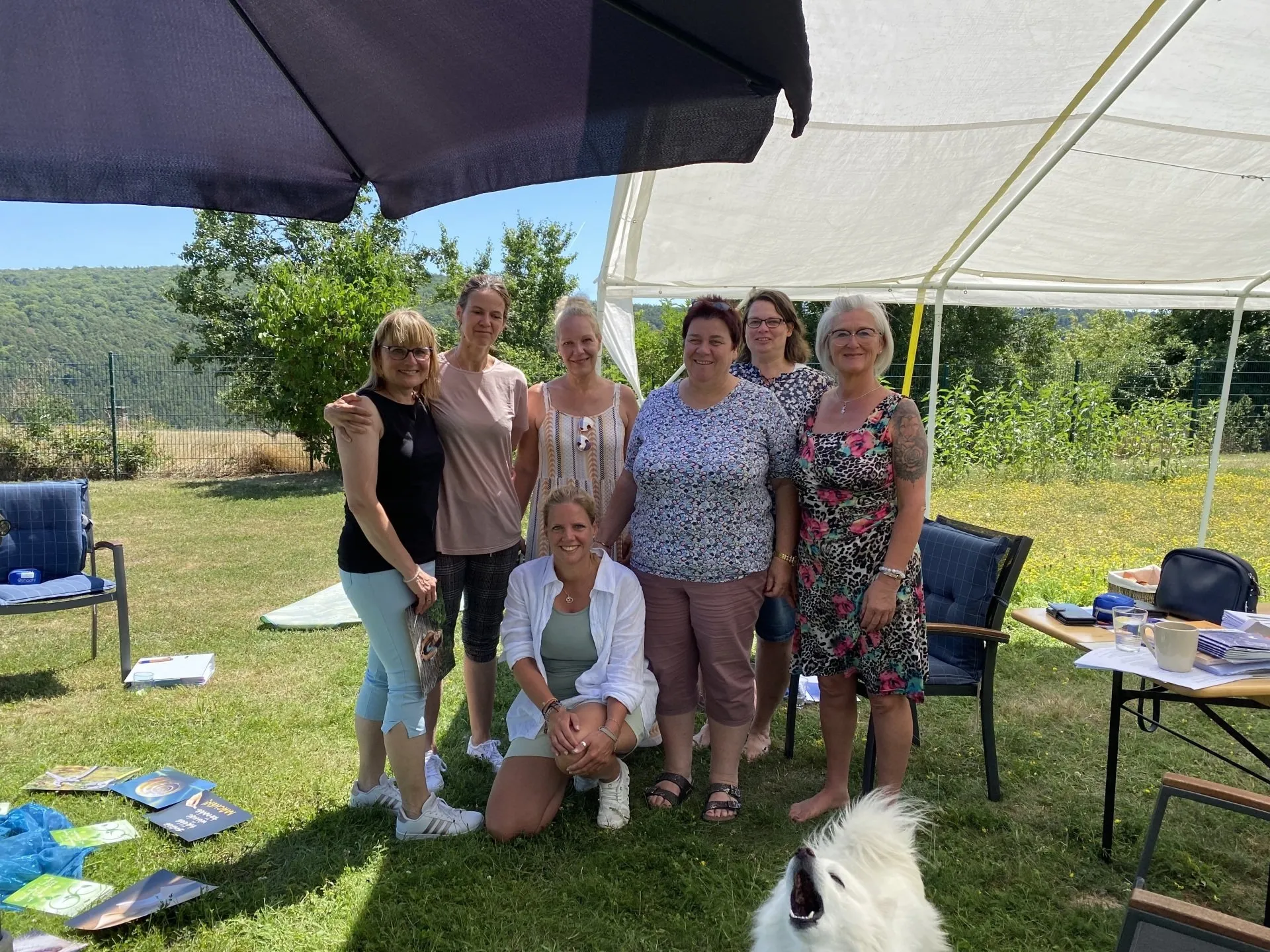 Eine Gruppe von 7 Frauen steht unter Sonnenschirmen auf einer Wiese und schaut glücklich in die Kamera.