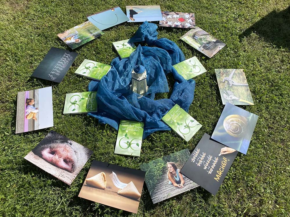 Kreis aus verschiedenen Postkarten und Bildern auf Gras, in der Mitte blaue Stoffbahnen und eine Laterne